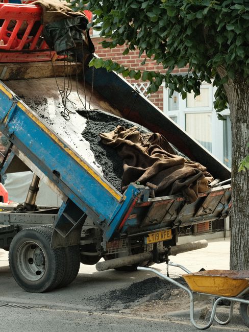 A blue and yellow construction dump truck parked on a residential street during daytime, with its bed tilted upward, unloading dark asphalt and a folded brown blanket or cloth. The truck is positioned near a tree with green foliage and a brick building with a window in the background. A red plastic crate is placed on top of the truck's cargo area, and a shovel with a yellow handle is on the ground nearby. The scene appears to be part of a road or pavement repair process, related to home or property maintenance, with possible involvement of a removal or transport service such as Мan with Van Norbiton.