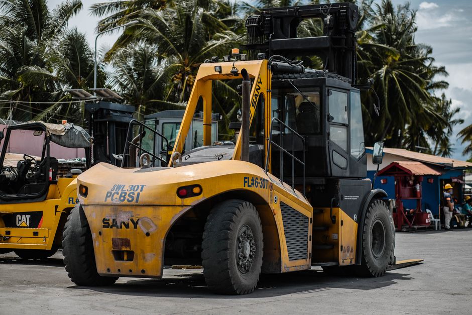 A yellow and black SANY forklift with large, heavy-duty tires is positioned outdoors on a paved surface, surrounded by other construction equipment including a smaller yellow Caterpillar vehicle. The forklift's cabin has glass windows, and the lifting mechanism is raised slightly above a variety of packed boxes and furniture covered with fabric or plastic wrap, which are arranged on the ground nearby. In the background, there are palm trees, a small building with a red roof, and a few workers wearing safety helmets, indicating a busy, outdoor environment typical for loading or unloading during a home relocation or construction site. The environment is well-lit with natural daylight, and the scene captures the machinery in the process of transporting or positioning items for a move or logistics operation, aligning with the themes of furniture transport and packing during house removals.