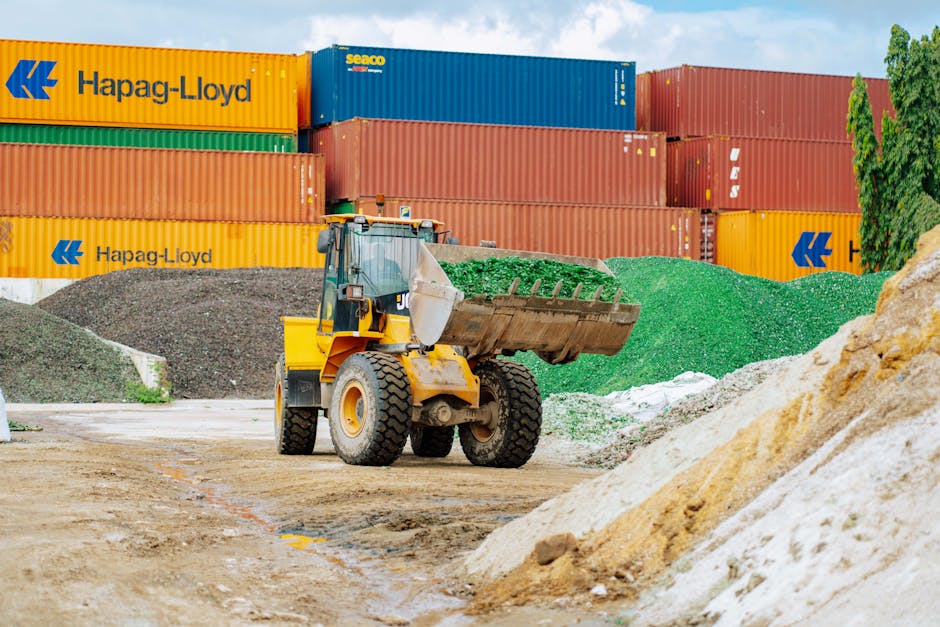 A blue and yellow construction dump truck parked on a residential street during daytime, with its bed tilted upward, unloading dark asphalt and a folded brown blanket or cloth. The truck is positioned near a tree with green foliage and a brick building with a window in the background. A red plastic crate is placed on top of the truck's cargo area, and a shovel with a yellow handle is on the ground nearby. The scene appears to be part of a road or pavement repair process, related to home or property maintenance, with possible involvement of a removal or transport service such as Мan with Van Norbiton.