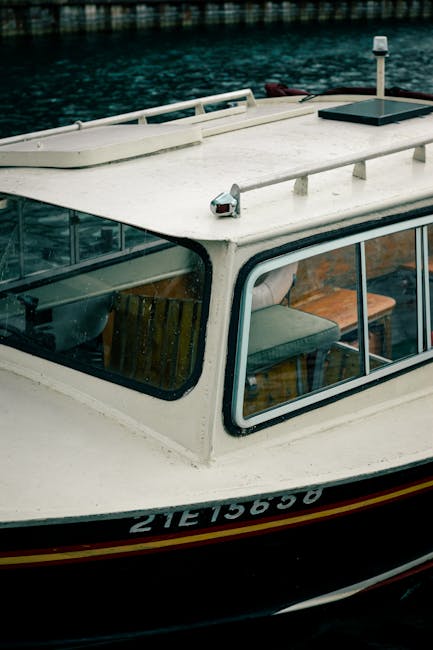 Close-up view of a boat docked on water, showing the white deck with a black stripe along the hull. The boat features large rectangular windows with black frames revealing wooden interior furniture including chairs and cushions. On the deck, there is a small black hatch and a white and grey navigation or communication device mounted on the roof. The boat appears to be in a calm water setting, with reflections visible on the surface, and is part of a house removal or transport process managed by Мan with Van Norbiton, specialising in removals and home relocation services, emphasizing the careful handling and transportation of domestic items during planned moves.