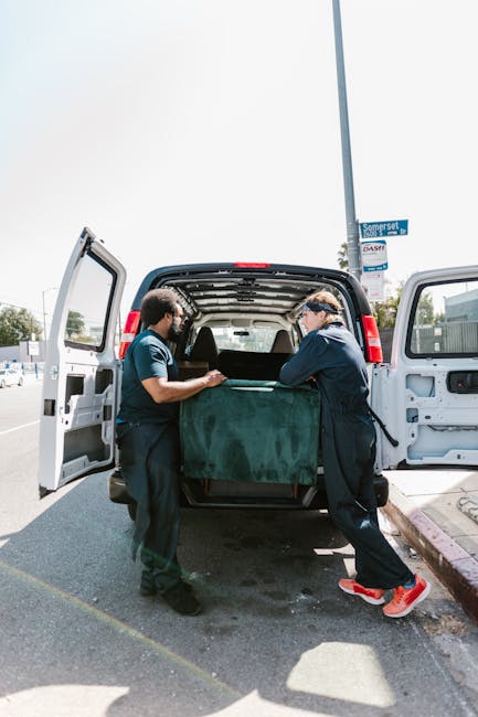Two movers from Мan with Van Norbiton are loading a large, dark green upholstered piece of furniture into the back of a white panel van during a home relocation in Kingston Hill. The van's rear doors are fully open, revealing the spacious interior with a black protective mat on the floor. The movers are wearing dark uniforms, and one is equipped with protective goggles, as they carefully lift and position the furniture inside. In the background, there are outdoor parking spaces, a tall streetlight, and a street sign indicating Dorset Road, suggesting the location is outside a residential or commercial property. The scene is lit with natural daylight, highlighting the urban surroundings and the detailed process of furniture transport, which is part of the broader packing and moving service provided by the company. This image captures the logistics involved in furniture transfer during a professional house or flat move, emphasizing the careful handling and organized loading process.