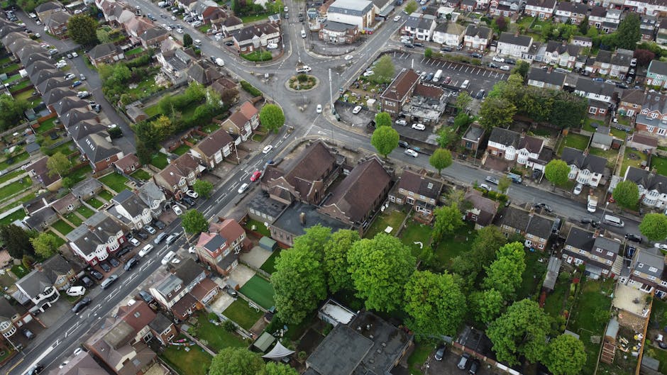 An aerial view of a residential neighbourhood showing densely packed houses, streets, and a small roundabout with a central garden feature. The area includes detached and semi-detached homes with varied roof styles, driveways, and parked cars. Several green trees are dispersed throughout the streets and gardens, providing greenery in the suburban landscape. The image captures the exterior of the houses and the surrounding environment, illustrating a typical urban area where house removals and furniture transport might take place, as managed by Мan with Van Norbiton. The scene is well-lit, with daylight illuminating the rooftops, streets, and foliage, emphasizing the individual property boundaries, layout, and accessibility for movers involved in property relocation and packing and moving services.