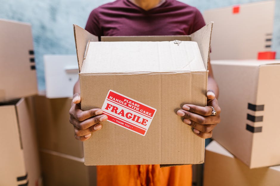 A person holding an open cardboard box with a red and white 'Fragile' handling sticker in a home relocation setting. The individual’s hands are visible, with neatly maintained nails and a ring on the left ring finger. The person is standing in a room filled with stacked cardboard boxes, some sealed with black packing tape, on a light-colored surface. Behind them, a portion of the person’s maroon shirt and orange trousers can be seen, with a blurred background that suggests an indoor space prepared for furniture transport. The boxes are of various sizes, some wrapped in plastic or fabric for protection during packaging and moving. The environment is well-lit, reflecting typical packing and moving processes managed by house removal specialists such as Мan with Van Norbiton. This scene illustrates the careful handling of fragile items during home relocation, aligning with services like packing and loading provided for moves from Penrhyn Road to Kingston in the KT2 Norbiton area.