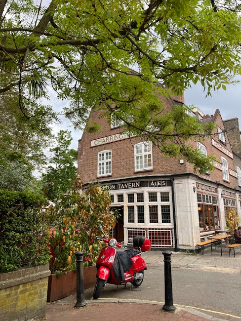 A red scooter parked on a paved sidewalk outside a traditional brick building with large front windows and signage indicating it's a pub or tavern, with outdoor seating visible on the right side. The scene is shaded by a large tree with green leaves extending across the top of the image, partially obscuring the building. The area appears to be part of a street or public space, with black metal bollards positioned along the edge of the pavement. The setting suggests a typical urban environment suitable for home relocation or moving services, with nearby space for loading or unloading furniture and boxes in a professional removal context, as operated by Мan with Van Norbiton.