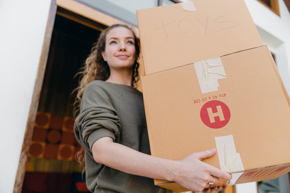 A young woman with curly hair, dressed in a grey long-sleeved top, stands indoors near a doorway, holding two cardboard boxes stacked one on top of the other. The top box is labeled with handwritten text 'toys', while the bottom box has a red and white label with a large 'H' symbol and printed dimensions. The boxes are secured with packing tape and appear to be filled with household items for a home relocation or moving process. The woman is smiling slightly and looking off into the distance, and natural light illuminates the scene. Inside the property, a patterned fabric or cushion is visible behind her, and the setting suggests a move-in or packing activity supported by professionals such as Мan with Van Norbiton, known for house removals and furniture transport. This image illustrates packing and moving preparations, emphasizing the handling of household goods during a house relocation or same-day move in Norbiton.
