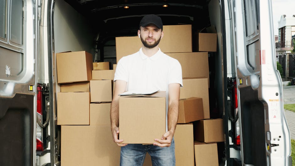 A man dressed in a white shirt, black cap, and jeans is standing at the open back of a moving van, holding a cardboard box during a home relocation process. The van's interior is filled with stacked c