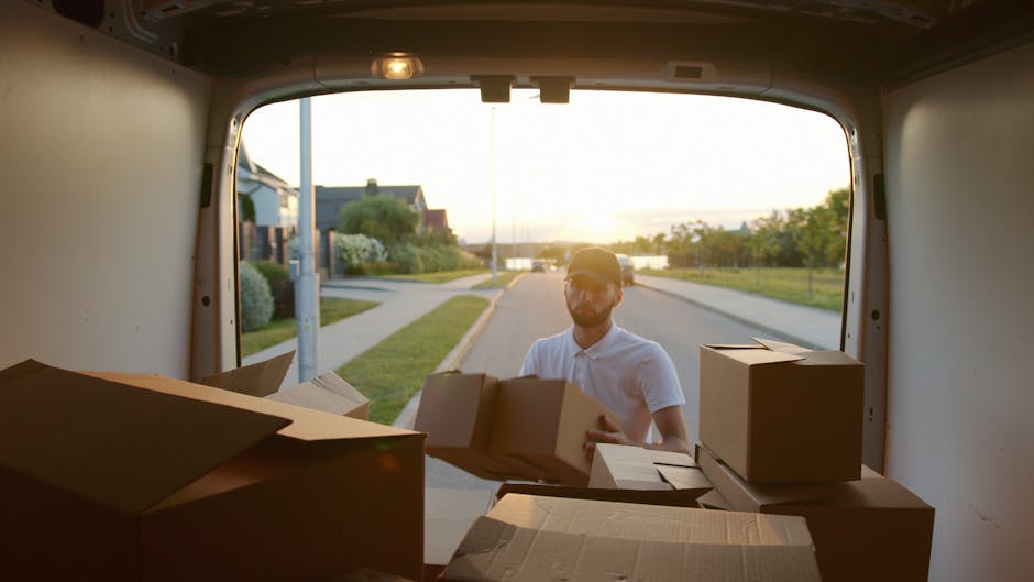 A man dressed in a white shirt, black cap, and jeans is standing at the open back of a moving van, holding a cardboard box during a home relocation process. The van's interior is filled with stacked c
