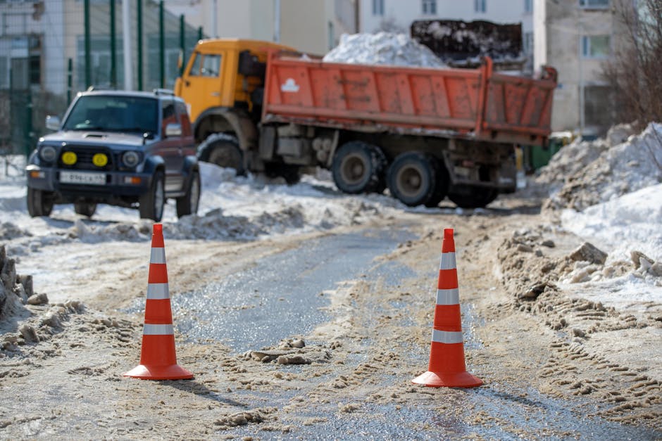 A gravel and snow-covered residential street area temporarily blocked off with two orange and white traffic cones placed on the uneven ground. Behind the cones, a yellow and black utility truck with a