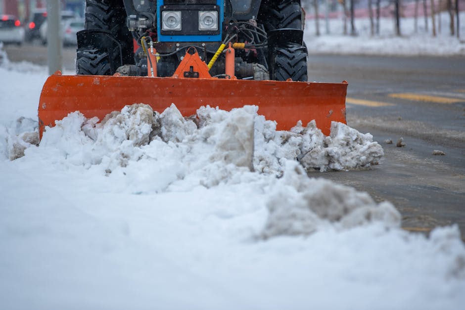 A gravel and snow-covered residential street area temporarily blocked off with two orange and white traffic cones placed on the uneven ground. Behind the cones, a yellow and black utility truck with a