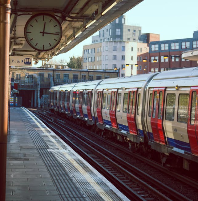 A London Underground train with red, silver, and blue exterior panels parked at Norbiton Station platform under an overhanging canopy. The platform has tactile paving along the edge and several closed doors on the train, indicating it is stationary. A large round clock hanging from the platform roof shows the time as approximately 2:10. Behind the train, modern multi-storey buildings with a mix of rectangular windows and brick or light-colored facades can be seen, with some trees in the background. The lighting suggests it is daytime, and the station is clean and orderly, prepared for a home relocation or moving process involving the transport of furniture and boxes. The scene is part of a typical London transport environment, relevant to services such as removals by Мan with Van Norbiton, emphasizing the importance of efficient moving logistics near Norbiton Station.