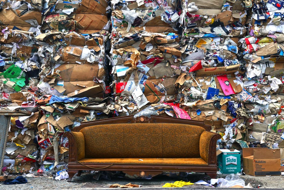 A vintage-style upholstered wooden sofa with a mustard-colored fabric seat is positioned on a sidewalk in front of a large, chaotic pile of mixed waste materials. The waste includes crumpled cardboard boxes, paper, plastic packaging, and torn paper bags, all haphazardly stacked and spilling onto the pavement behind the sofa. Some items are partially covered with plastic wrap or tied with string, and a few cardboard boxes are visible at the base of the pile. The scene suggests a setting related to bulk waste disposal or clearance, with the sofa contrasting against the clutter of household and recyclable waste. The background is filled with tangled, compressed waste materials, indicating a location where large amounts of waste have been accumulated or disposed of, possibly related to house removals or furniture disposal services, as referenced by the website manwithvannorbiton.co.uk.