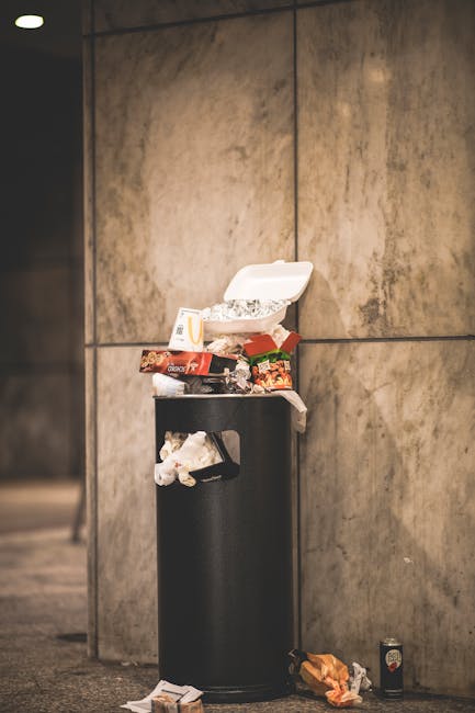 A large green wheelie bin positioned outdoors on a paved surface next to a dark brick wall, filled with red and black packaging bags labeled 'FK200 Flexy Plus' and other flex hose accessories. The bin is partially open, revealing the contents, which include cardboard boxes and various plastic packaging materials. The bin is mounted on wheels, with a white label displaying contact details for a disposal service, and is situated adjacent to another slightly smaller green bin. Items such as a metal ramp or step and a cardboard piece are placed on the ground nearby. This scene reflects a typical home removal or waste disposal process, involving packing materials and collection for recycling or disposal, relevant to house relocations or furniture transport tasks conducted by companies like Мan with Van Norbiton.