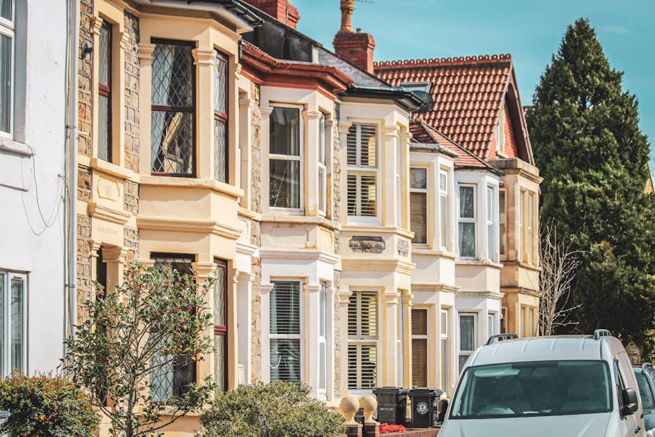 A row of traditional Victorian-style terraced houses with ornate bay windows, decorative stonework, and cream-colored painted facades, situated on a residential street in Norbiton. The houses are three stories high, with red-tiled pitched roofs, and surrounded by greenery including trees and shrubs. In the foreground, a white van belonging to Мan with Van Norbiton is parked on the pavement, partially visible. The van is positioned near the entrance to one of the properties, where a systematic home relocation or furniture transport process is taking place. The scene captures the exterior environment during daylight, with clear blue skies overhead, and the setting suggests a busy but organized move-in or move-out activity involving packing supplies, such as cardboard boxes and fabric wrapping, and the use of moving equipment like trolleys, as part of a professional removals service specializing in protecting period features during moves.