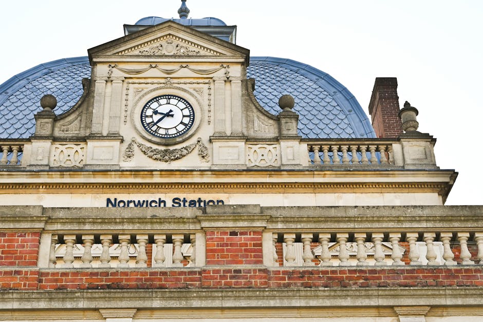 Close-up view of the upper section of Norwich Station's historic building facade, featuring a large round clock with black hands and Roman numerals set within an ornate stone frame. The building's architectural details include decorative stonework, balustrades, and rounded stone finials on either side of the roofline. The roof is clad in blue slate tiles, with a chimney visible on the right side. Below the clock, there is a sign reading 'Norwich Station' mounted on the stone ledge, with visible red brickwork and cream-colored stone balustrades surrounding the roofline. This image captures the exterior of the station during daylight, with natural lighting highlighting the intricate stone and brick detailing, providing context for house removals and furniture transport related to residential relocations involving Norwich Station.