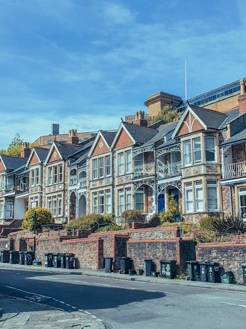 A row of traditional Victorian-style terraced houses with ornate bay windows, decorative stonework, and cream-colored painted facades, situated on a residential street in Norbiton. The houses are three stories high, with red-tiled pitched roofs, and surrounded by greenery including trees and shrubs. In the foreground, a white van belonging to Мan with Van Norbiton is parked on the pavement, partially visible. The van is positioned near the entrance to one of the properties, where a systematic home relocation or furniture transport process is taking place. The scene captures the exterior environment during daylight, with clear blue skies overhead, and the setting suggests a busy but organized move-in or move-out activity involving packing supplies, such as cardboard boxes and fabric wrapping, and the use of moving equipment like trolleys, as part of a professional removals service specializing in protecting period features during moves.
