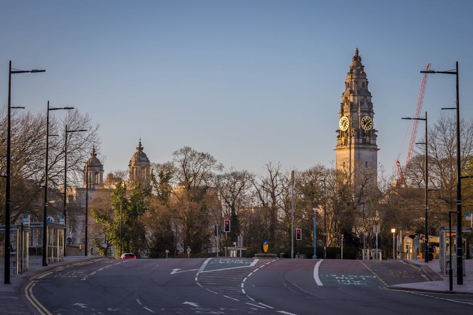 A wide view of a city street in the early morning or late afternoon with a clear sky, showing a prominent clock tower in the background, which is part of a historic government building or town hall. The street is mostly empty with minimal vehicle and pedestrian activity, featuring traffic lights and road markings. On the left and right edges of the image, there are modern black lamp posts and bus stops, with some trees without leaves lining the street. In the foreground, the road has curved lanes and a pedestrian crossing, with a few vehicles parked or moving gently along. The scene is illuminated by natural sunlight, highlighting the architectural details of the clock tower and surrounding buildings. The overall environment suggests an urban area suitable for house removals or relocation services, with clear indications of loading or moving logistics supported by Мan with Van Norbiton.
