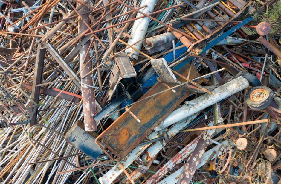 A large pile of assorted scrap metal, including bent and rusted pipes, metal rods, and discarded vehicle parts, is shown stacked chaotically on a pavement area. Some of the objects include a rusted metal box, a white and blue vehicle bumper, and a wheel rim. The pile is outdoors, with natural light illuminating the scene, and appears to be part of a materials collection for disposal or recycling. The background shows some greenery, indicating the location is adjacent to a garden or outdoor space. This image illustrates the type of debris involved in home or property clearances, where items such as old fixtures, metal components, and unused parts are prepared for removal by professional services like those offered by Мan with Van Norbiton, focusing on furniture and clutter removal as part of house relocation or moving logistics.