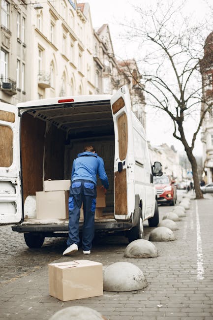 A man wearing a blue jacket and dark trousers is seen loading a large cardboard box into the back of a white delivery van parked on a city street. The van's rear doors are open, revealing the interior with visible wooden paneling and some packed items. Several cardboard boxes are placed on the pavement nearby, ready for transportation as part of home relocation or furniture transport. The street is lined with multi-storey residential buildings and a leafless tree, with a few parked cars visible further down the road. The scene is set in daylight, with natural light illuminating the area, demonstrating the loading process involved in professional removals services such as those offered by Мan with Van Norbiton.