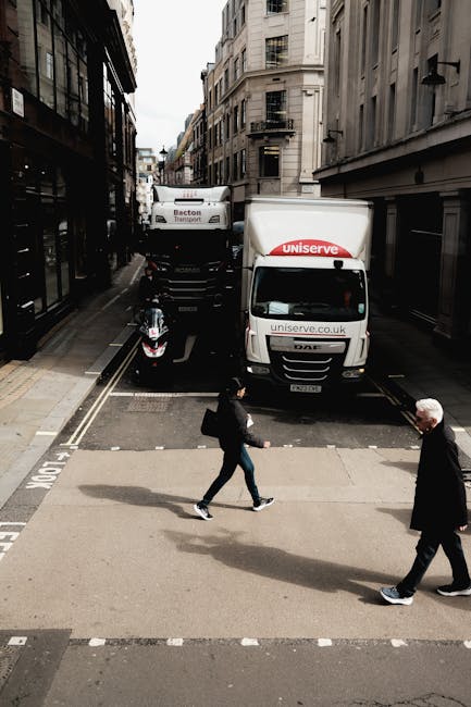 A street scene capturing the loading process of a house removal in Norbiton, where two delivery vans branded with 'uniserve.co.uk' and 'Becton Transport' are parked side by side on a narrow urban road. The vans are positioned directly adjacent to the building's entrance, with one partially over the curb and the other on the street. In front of the vehicles, a woman dressed in dark clothing is walking across the pedestrian crossing, carrying a bag, while an elderly man with white hair in a dark coat follows behind her, also crossing the road. The street is lined with tall, multi-story buildings with a mix of modern and older architectural styles, and the weather appears overcast, with diffuse natural light illuminating the scene. The image illustrates the logistics involved in home relocation, highlighting the transportation of furniture and packing materials via professional moving services such as those offered by Мan with Van Norbiton, focusing on the loading process and street-side preparation for a house removal.